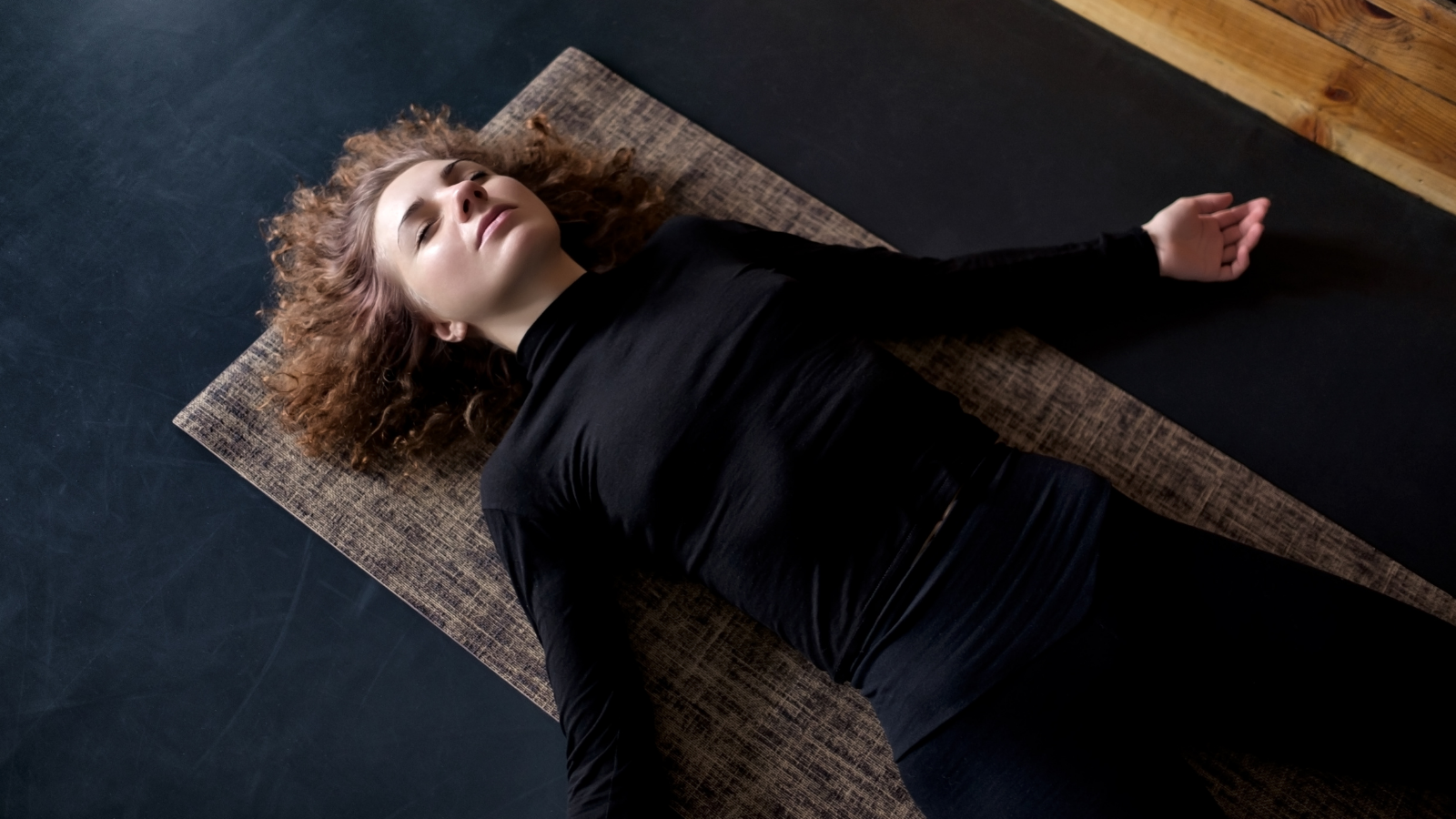 Yoga's Savasana can be a great way to prepare your mind nd body for a great night's sleep Young curly woman practicing in a yoga studio resting in Savasana or Corpse Pose at the end of practice.