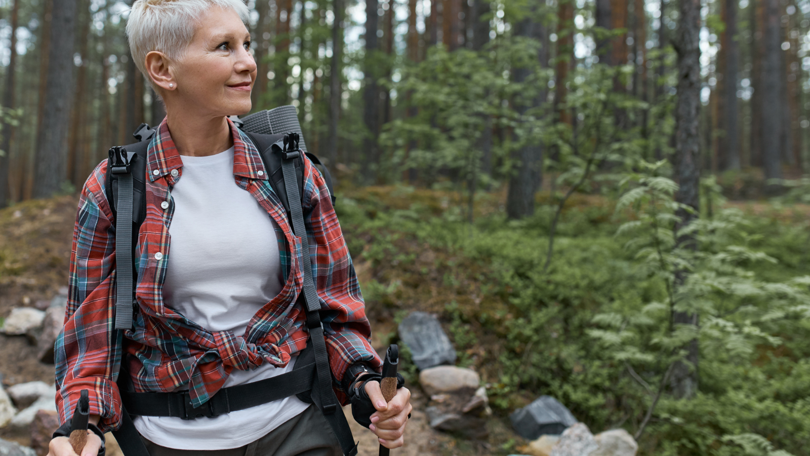 Aging, people, active lifestyle and health concept. Outdoor portrait of happy European female walker with backpack and poles, enjoying beautiful nature while nordic walking in pine forest.