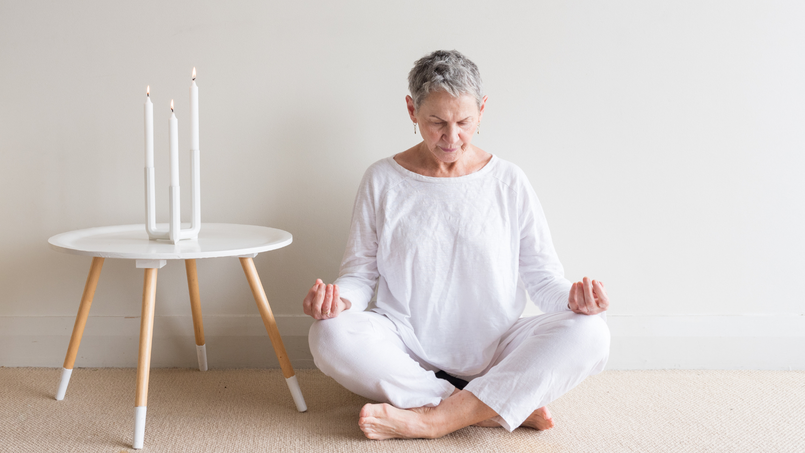 How to practice two to one breathing. Beautiful older woman in white clothing sitting in yoga meditation position with candles against neutral wall background